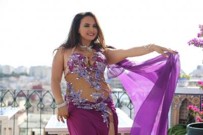 Femme en tenue de danse orientale violette ornée de motifs brillants, posant devant des fleurs et un ciel dégagé.