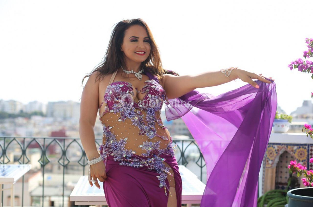 Femme en tenue de danse orientale violette ornée de perles, posant sur une terrasse ensoleillée avec vue urbaine.