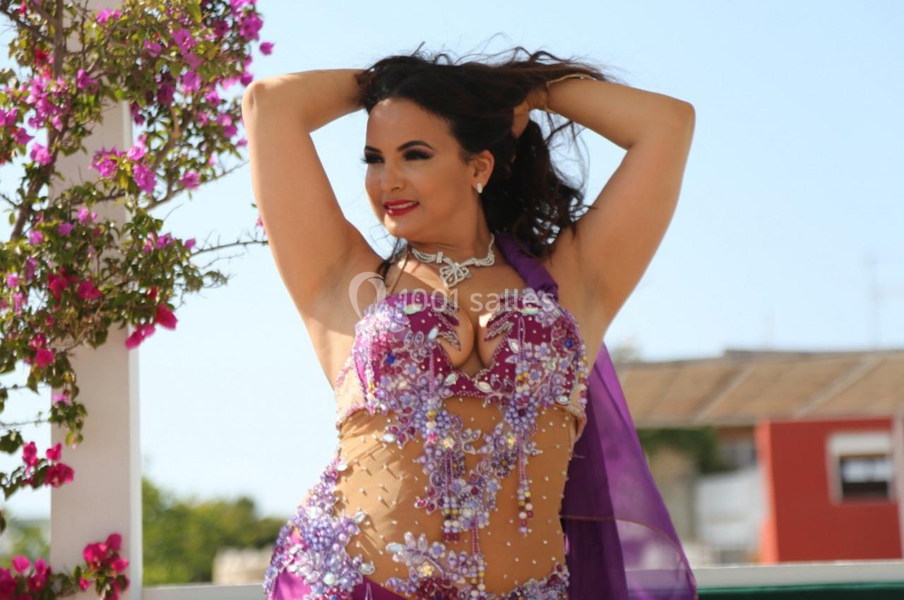 Femme en tenue de danse orientale violette ornée de perles, posant devant un fond extérieur fleuri et ensoleillé.
