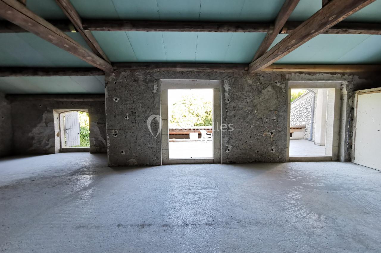 Intérieur d'une pièce vide avec murs en pierre, plafond en bois et ouvertures donnant sur un extérieur lumineux.