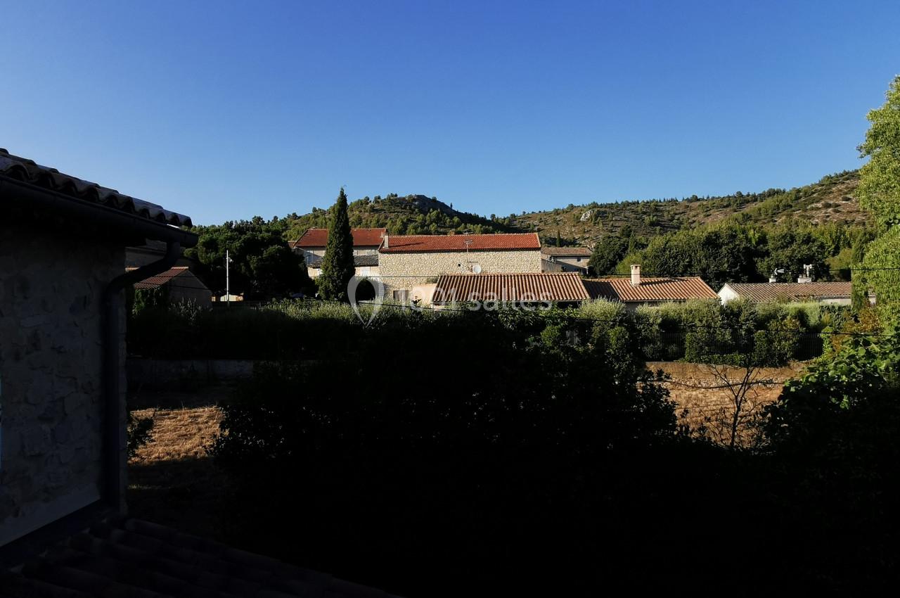 Vue d'un village avec des toits en tuiles rouges, entouré de végétation et de collines sous un ciel bleu clair.