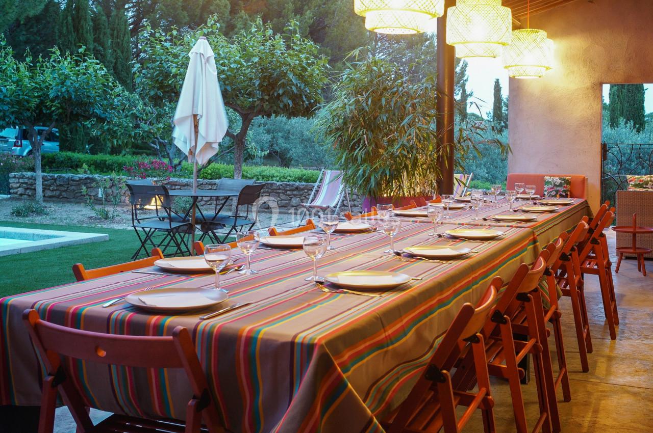 Grande table dressée avec une nappe colorée sous une terrasse éclairée, donnant sur un jardin avec piscine.