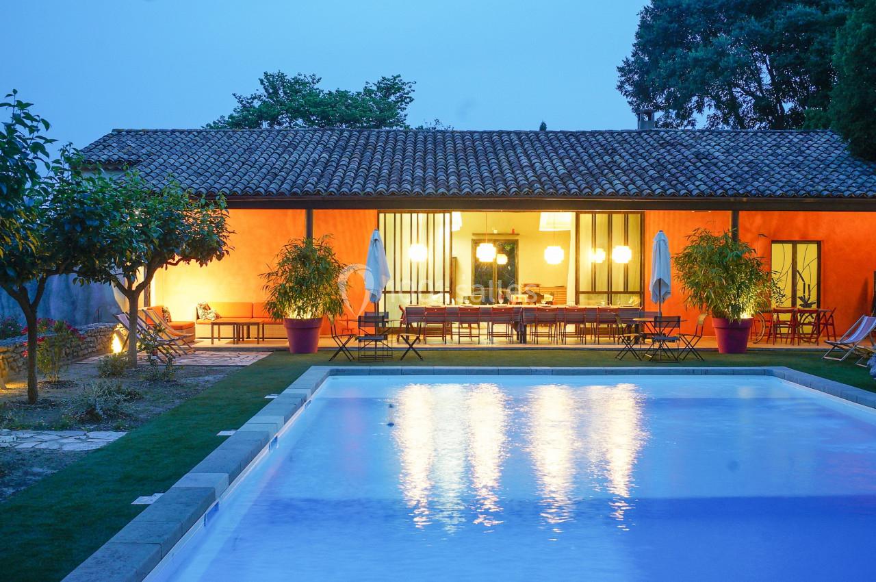 Maison avec façade orange, éclairée en soirée, entourée de végétation, avec une piscine au premier plan.