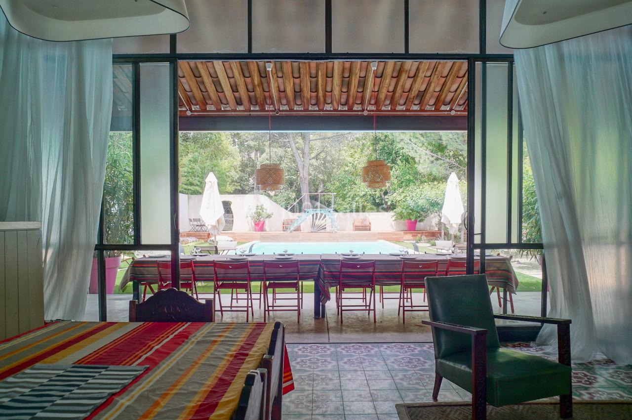 Vue sur une terrasse aménagée avec une grande table, donnant sur une piscine entourée de verdure et de parasols blancs.