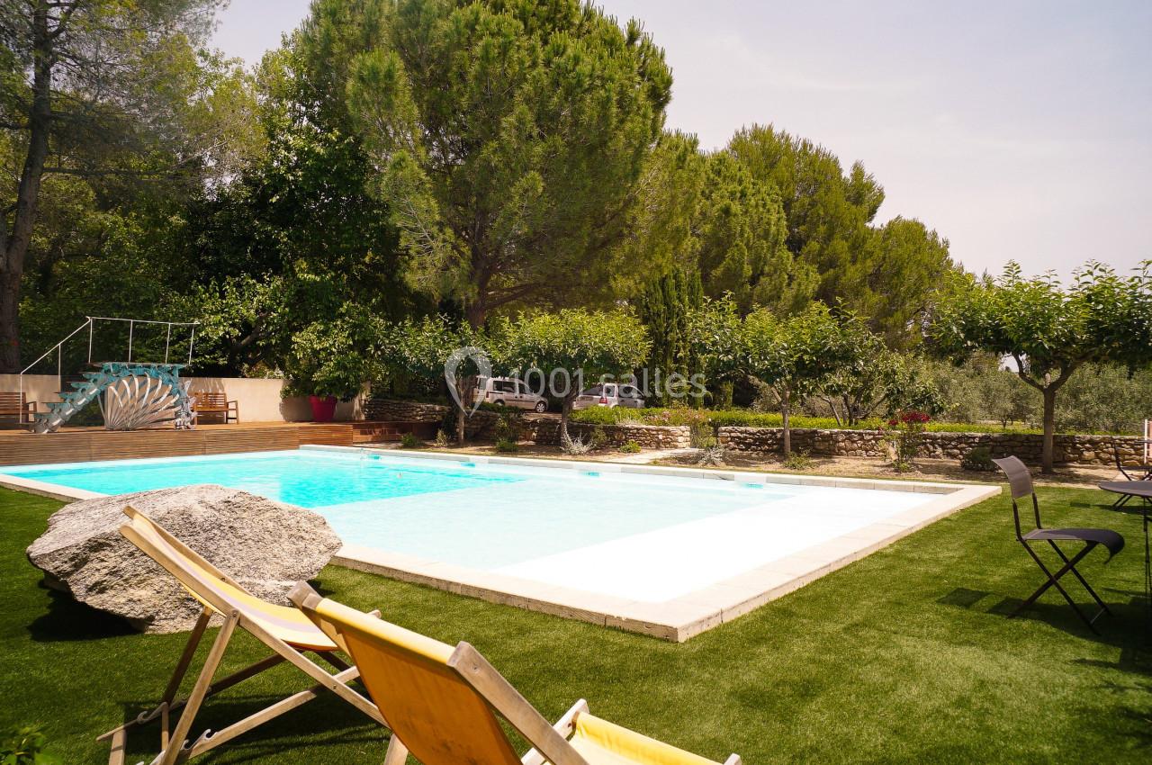 Piscine extérieure entourée de verdure, avec des chaises longues et des arbres sous un ciel dégagé.