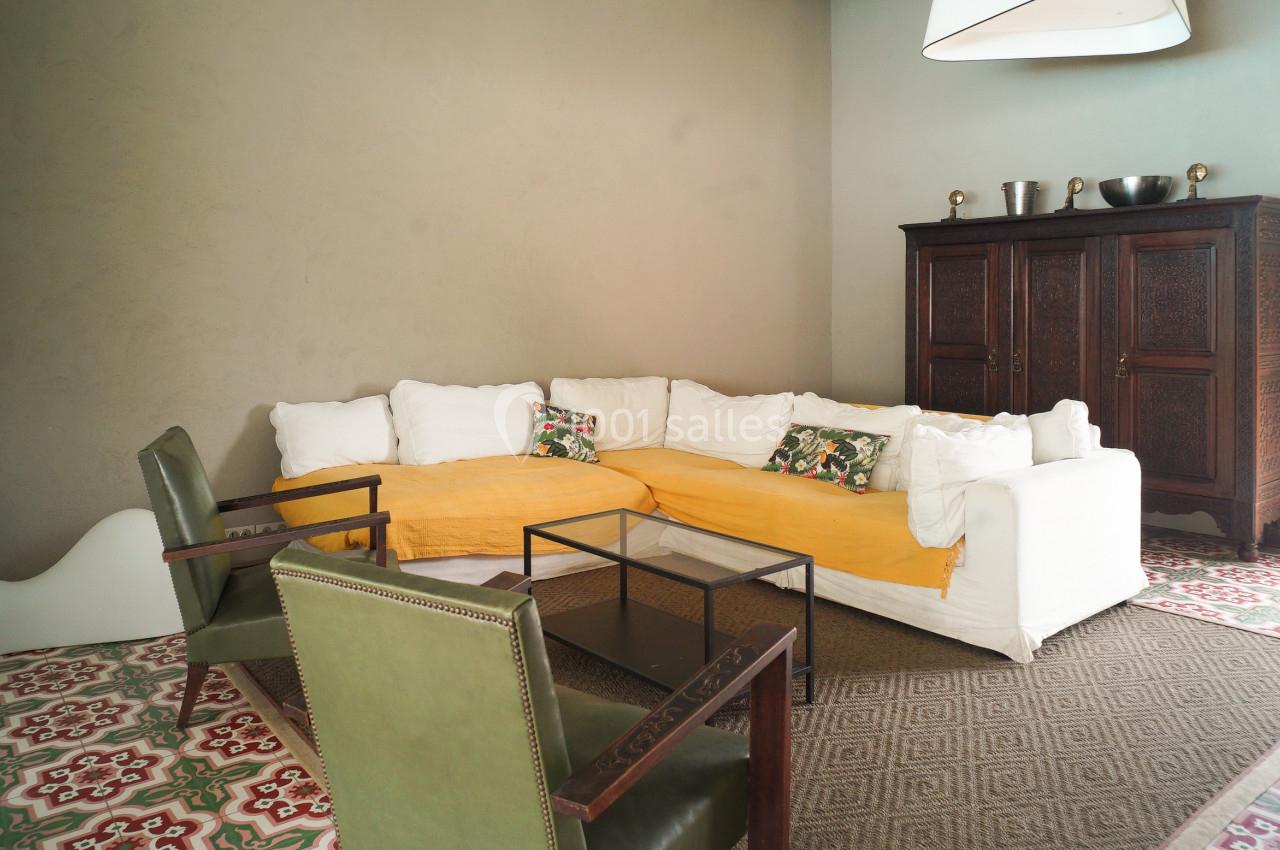 Salon avec canapé d'angle blanc, plaid jaune, fauteuils verts, armoire en bois sombre et carrelage coloré au sol.