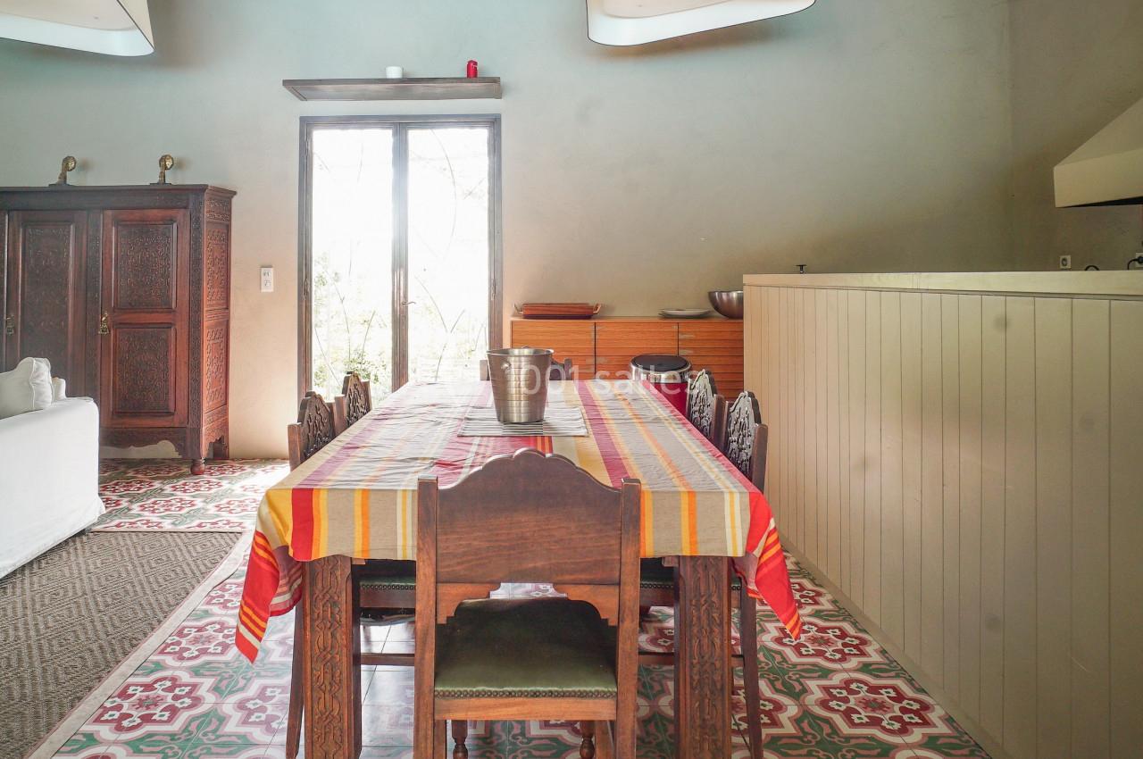 Salle à manger lumineuse avec une grande table recouverte d'une nappe colorée, entourée de chaises en bois.