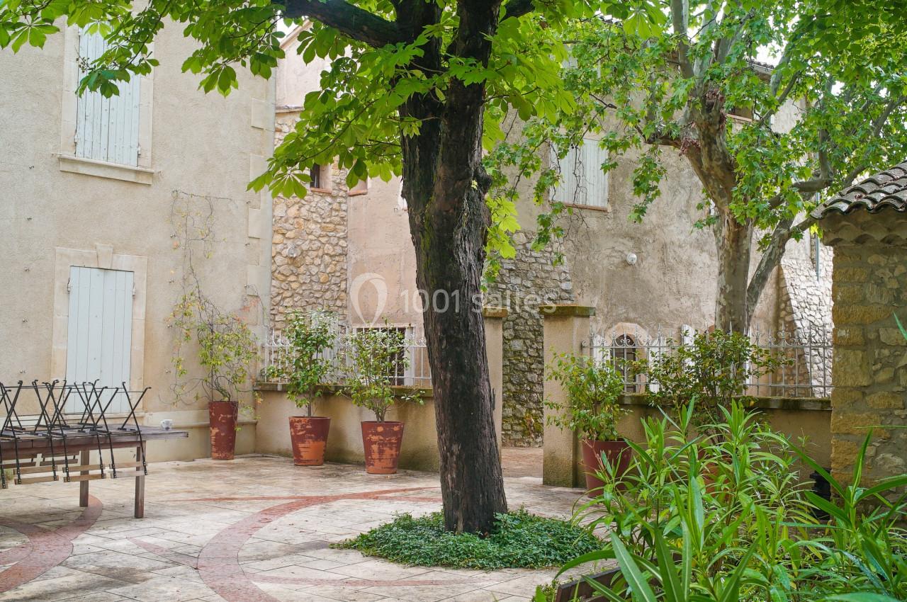 Cour intérieure en pierre avec un arbre central, des plantes en pots, des volets fermés et une table en métal.