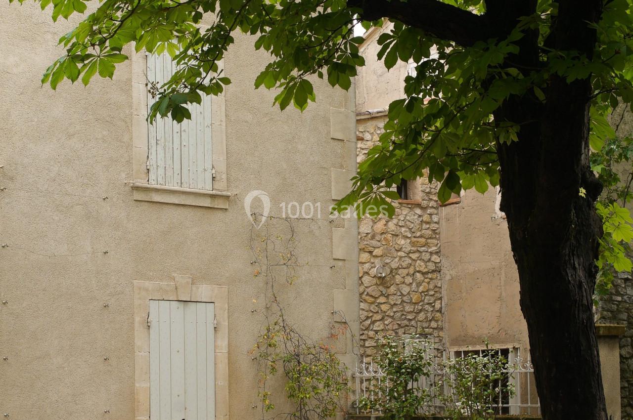 Cour pavée avec un arbre, des chaises empilées, des pots de fleurs et une façade beige aux volets fermés.