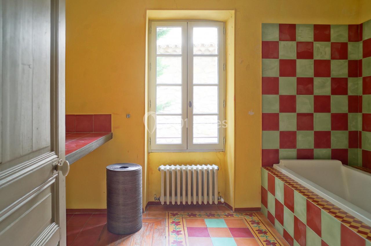 Salle de bain lumineuse avec murs jaunes, baignoire à carreaux rouges et verts, et fenêtre donnant sur l'extérieur.