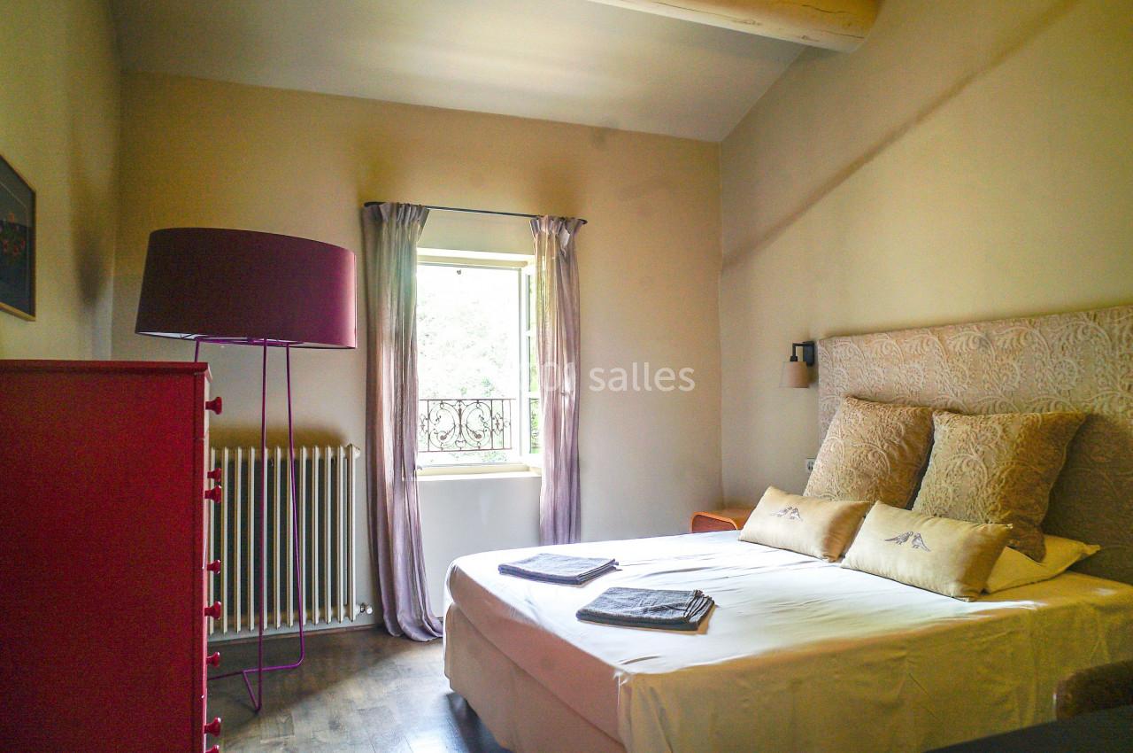 Chambre lumineuse avec lit double, coussins décoratifs, commode rouge, lampe sur pied et fenêtre donnant sur l'extérieur.