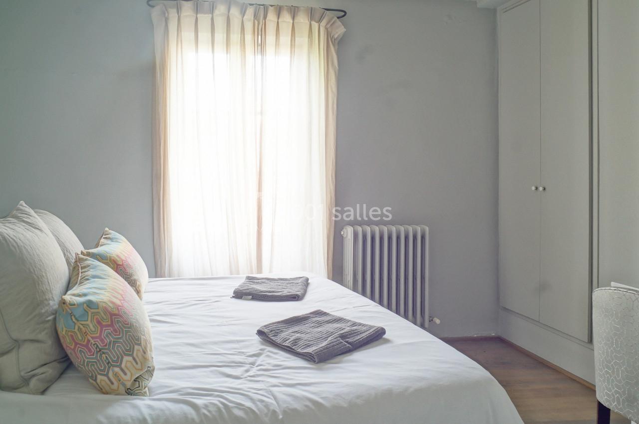 Chambre lumineuse avec lit double, coussins colorés, armoire blanche, radiateur et fenêtre avec rideaux clairs.