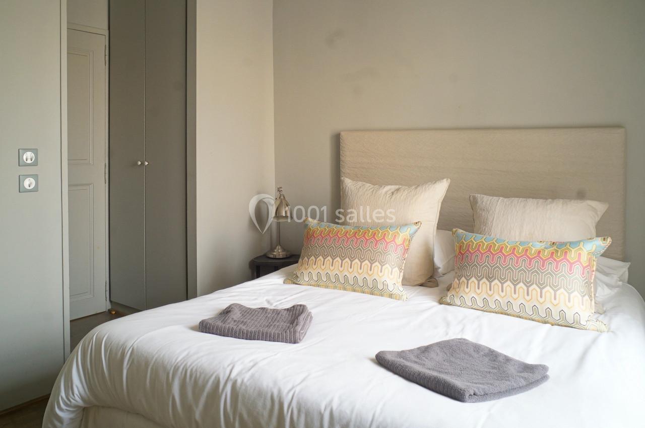 Lit double avec coussins colorés et serviettes pliées, dans une chambre lumineuse aux murs clairs et mobilier minimaliste.