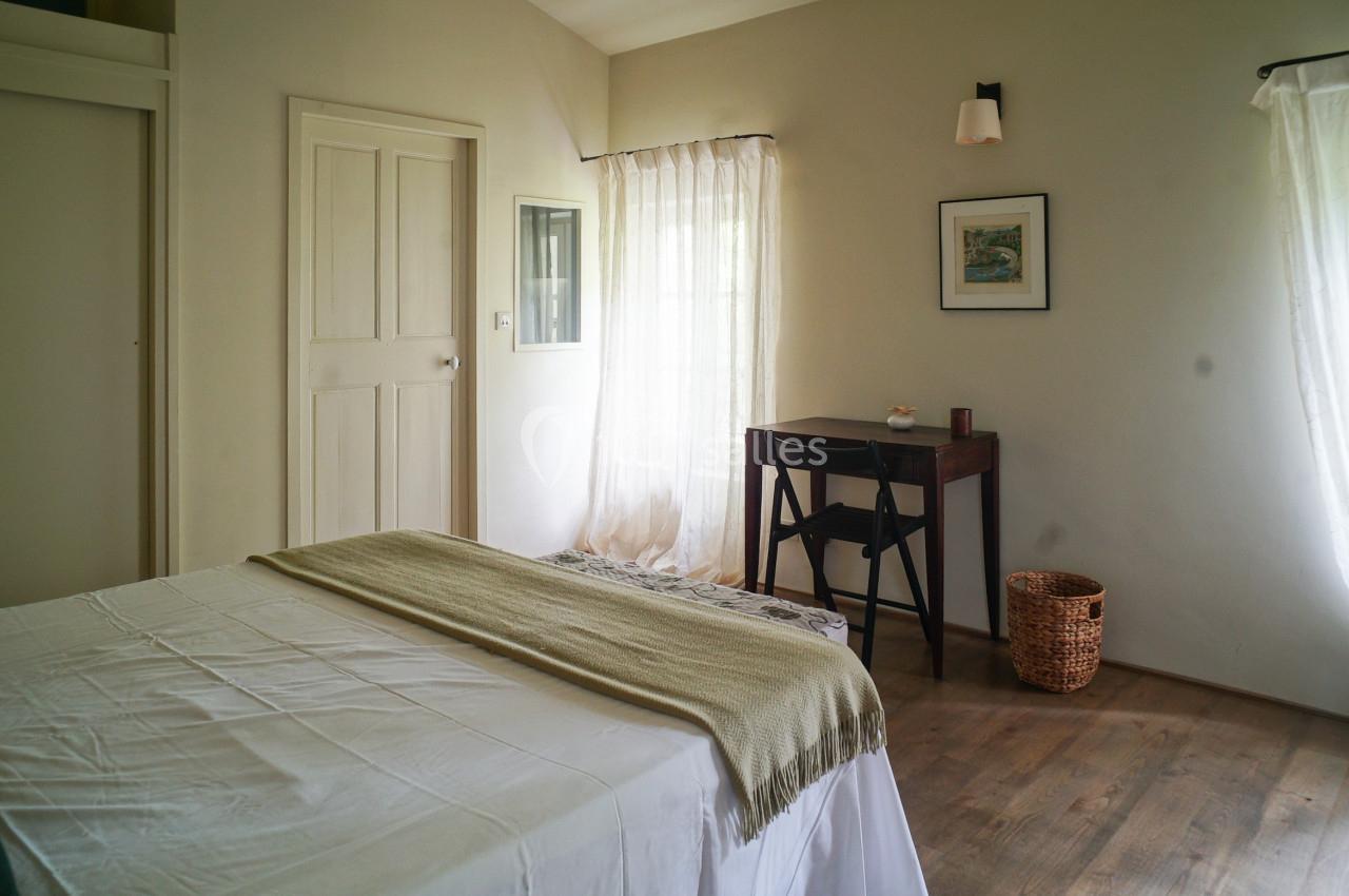 Chambre lumineuse avec lit double, bureau en bois, panier en osier et rideaux blancs laissant passer la lumière naturelle.