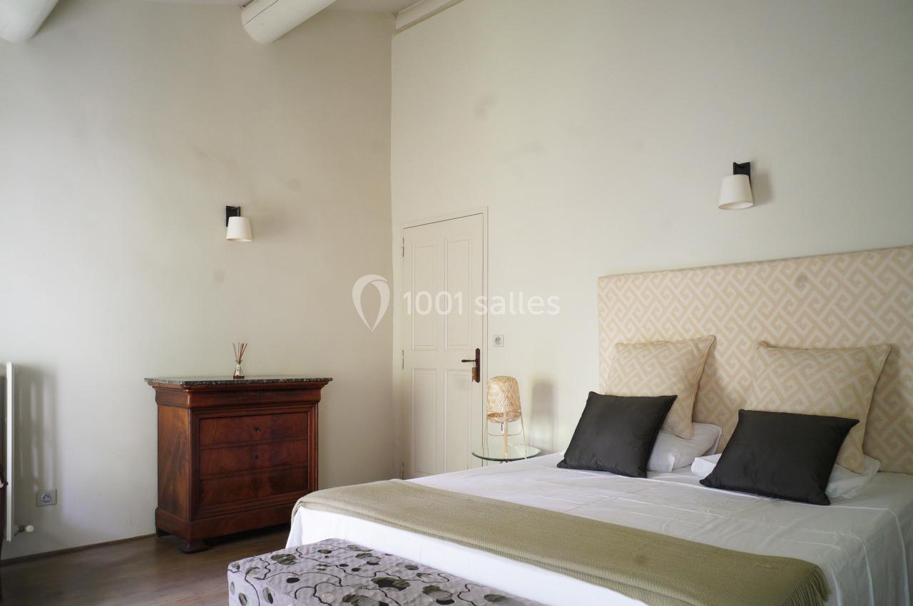 Chambre lumineuse avec lit double, tête de lit beige, commode en bois, chaise et murs blancs épurés.