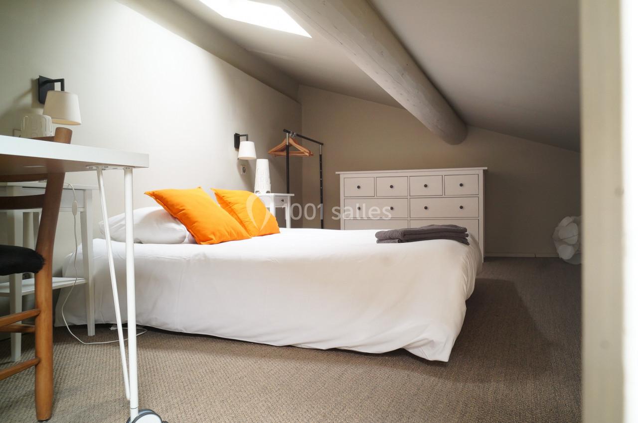 Chambre mansardée lumineuse avec lit double, coussins orange, commode blanche et bureau en bois clair.