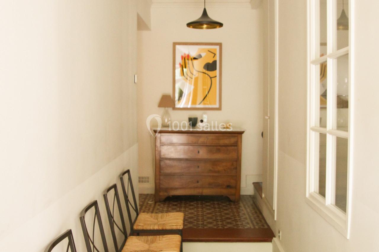 Couloir étroit avec carrelage géométrique, chaises alignées à gauche, commode en bois et tableau au fond.