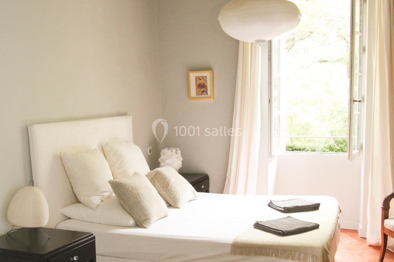 Chambre lumineuse avec lit double, tables de chevet noires, lampe suspendue et fenêtre ouverte sur un jardin.