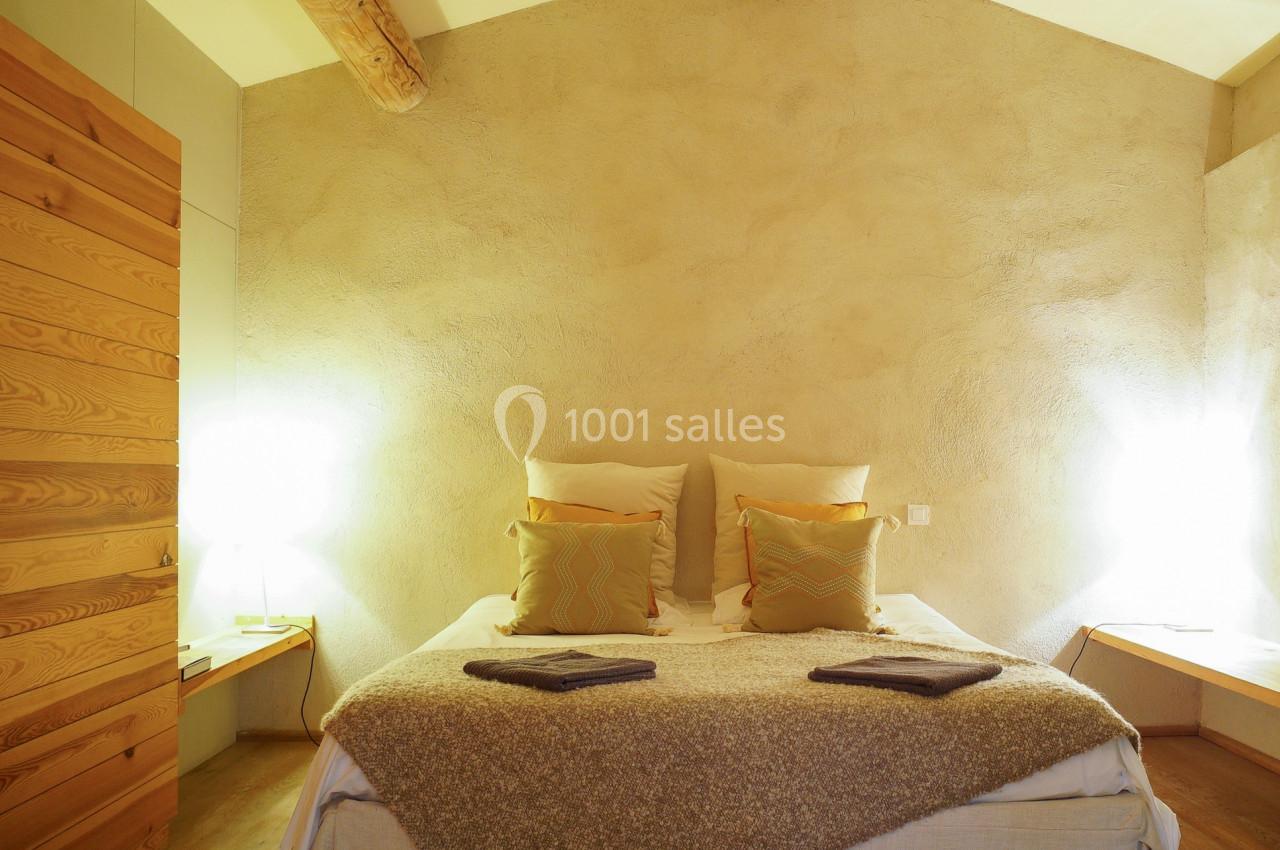 Chambre avec lit double, murs en enduit beige, poutres apparentes et éclairage doux sur des tables de chevet.