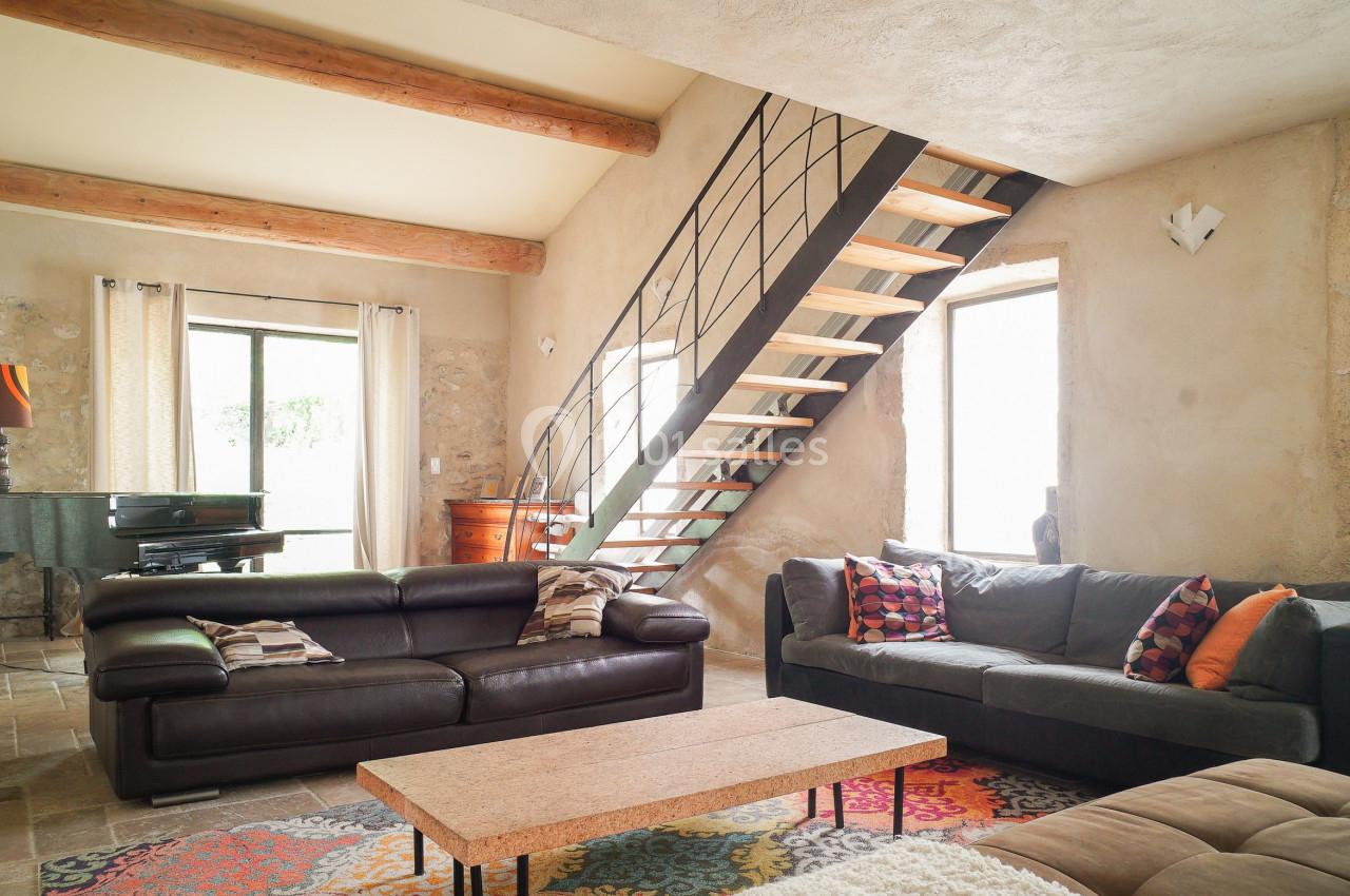 Salon lumineux avec murs en pierre, canapé en cuir, tapis coloré, escalier en métal et piano près d'une fenêtre.