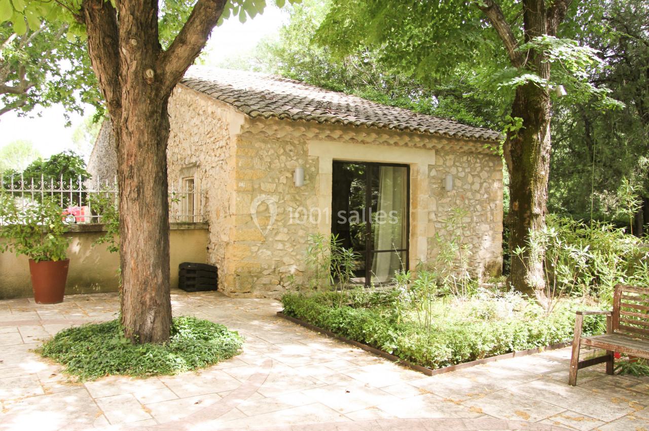 Petite maison en pierre entourée de verdure, avec une terrasse pavée ombragée par des arbres.
