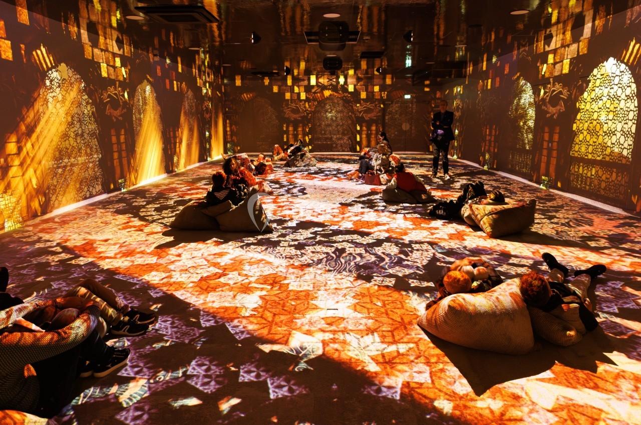 Salle immersive avec des projections lumineuses colorées sur les murs et le sol, des personnes assises sur des poufs.