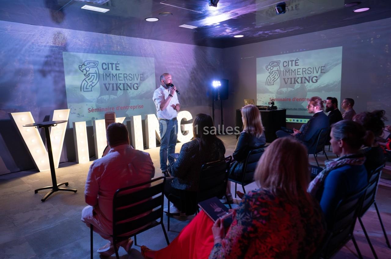 Un conférencier s'adresse à un public assis lors d'un séminaire d'entreprise, avec des projections et des lettres lumineuses.