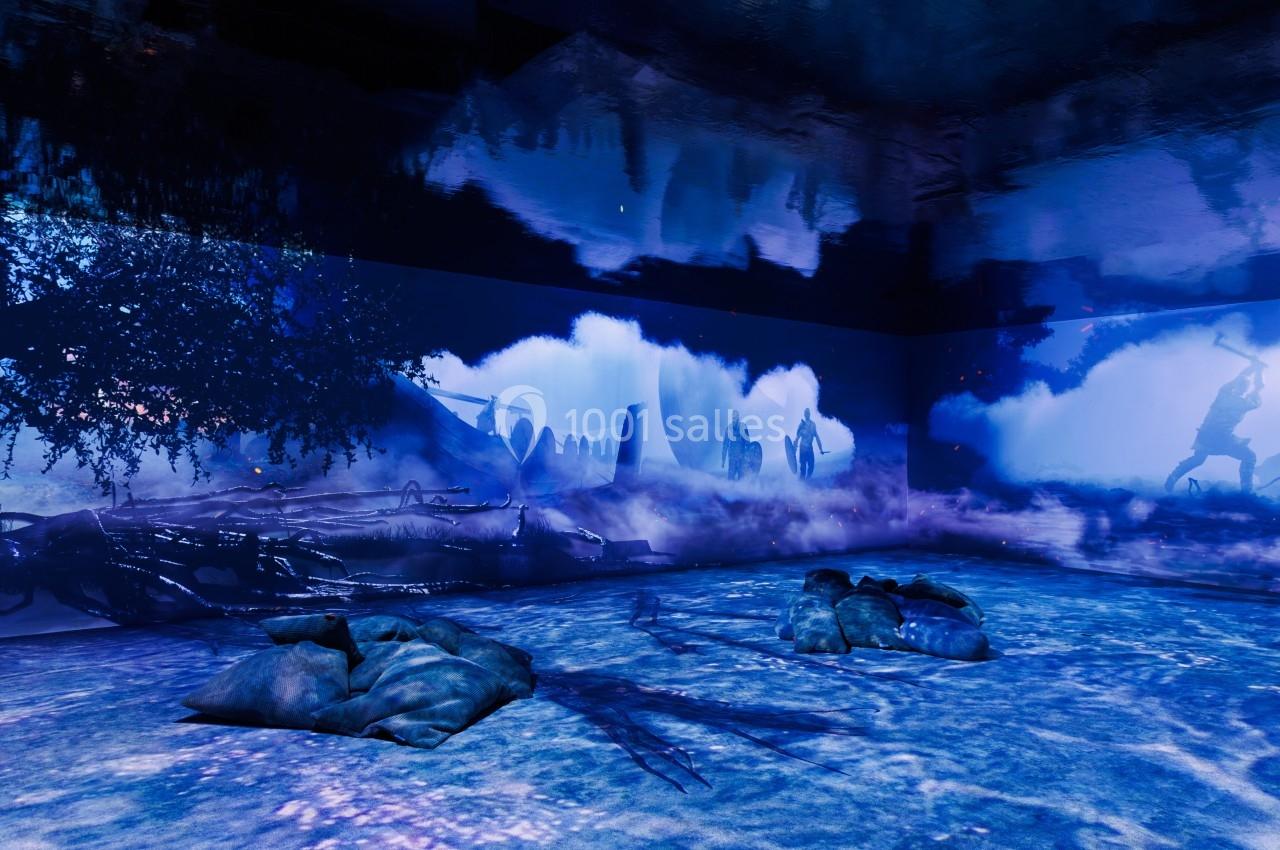 Projection immersive avec des paysages bleutés, des silhouettes humaines et des sacs posés au sol dans une salle sombre.