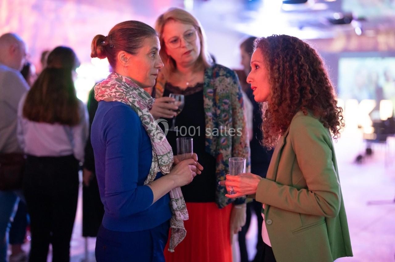 Trois femmes discutent debout dans un espace intérieur éclairé, tenant des verres à la main.