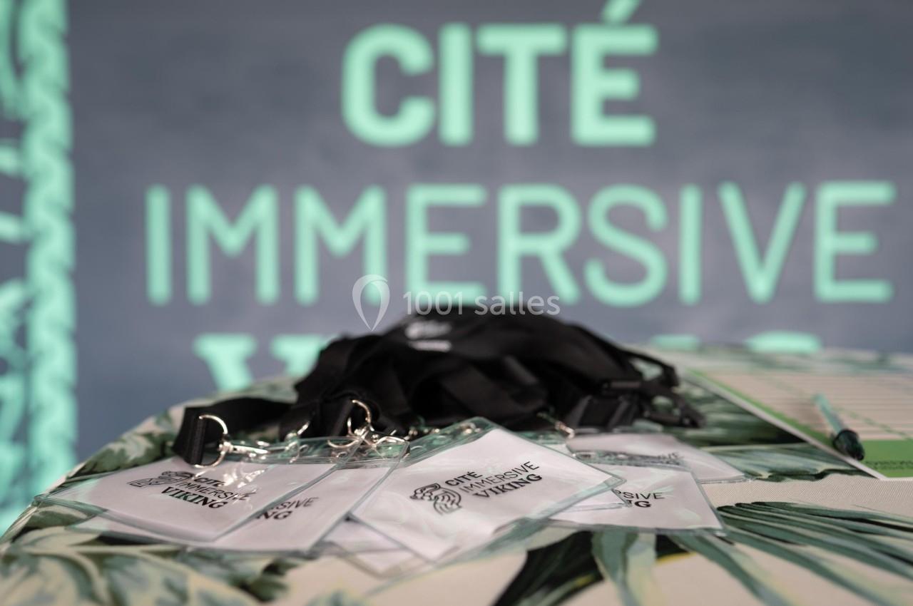 Des badges avec cordons noirs posés sur une table, devant un fond flou affichant ’Cité Immersive Viking’.