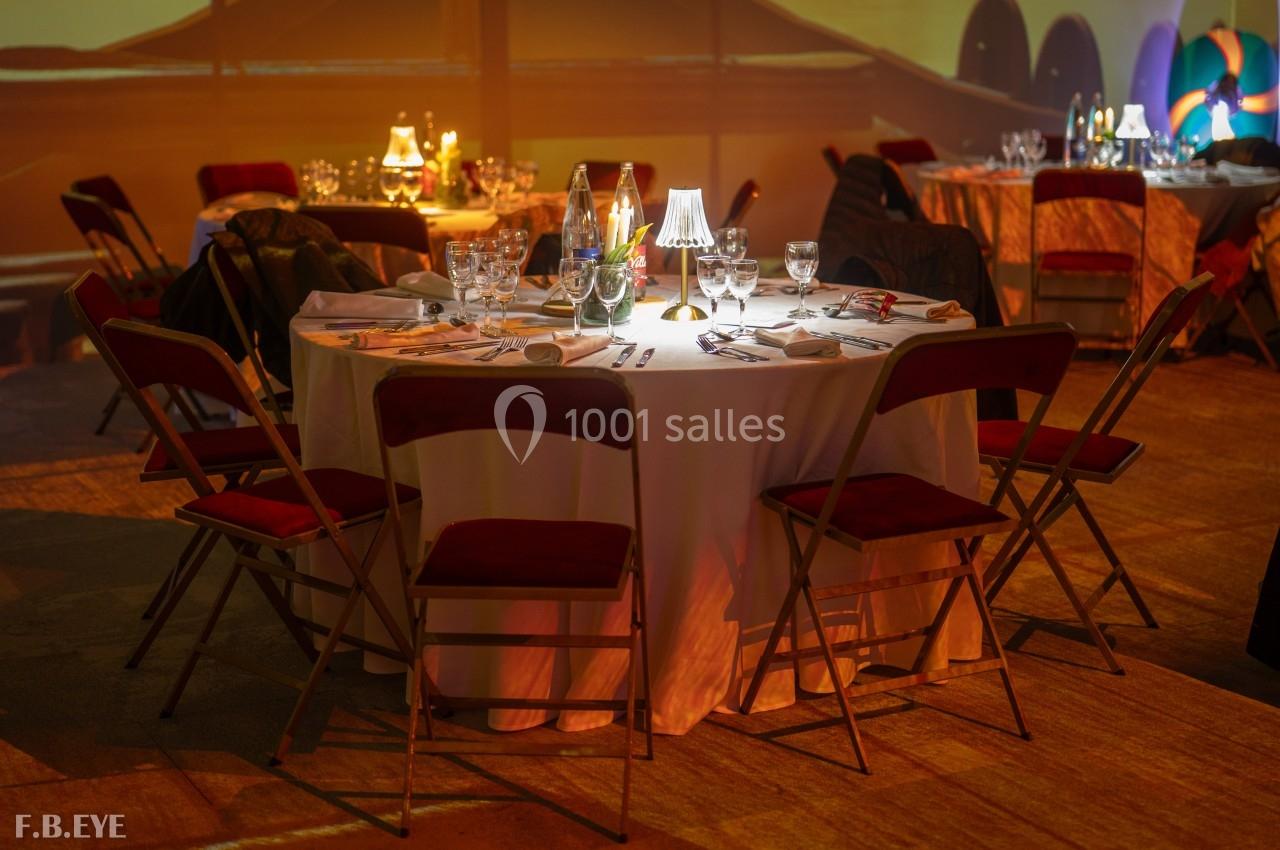 Table ronde dressée avec nappes blanches, couverts, verres et petite lampe, entourée de chaises rouges dans une salle…