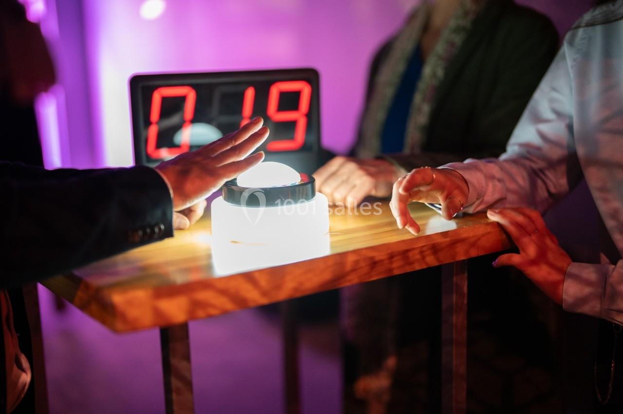 Des mains s'apprêtent à appuyer sur un buzzer lumineux posé sur une table, avec un écran affichant ’019’ en arrière-plan.