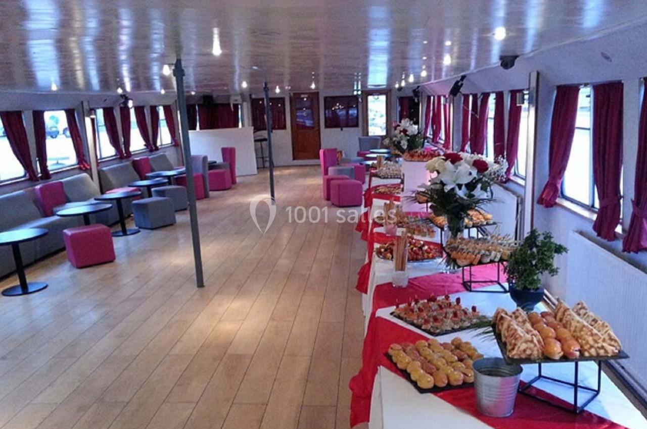 Salle de réception sur un bateau avec buffet dressé, comprenant des pâtisseries et amuse-bouches sur des nappes rouges.