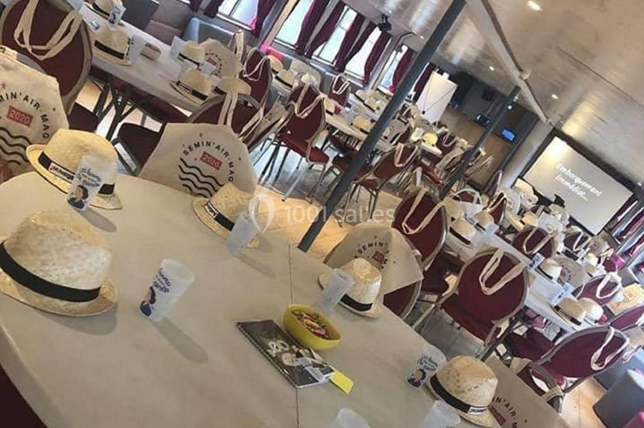 Salle aménagée avec des tables couvertes de chapeaux en paille, gobelets et sacs, prête pour un événement.