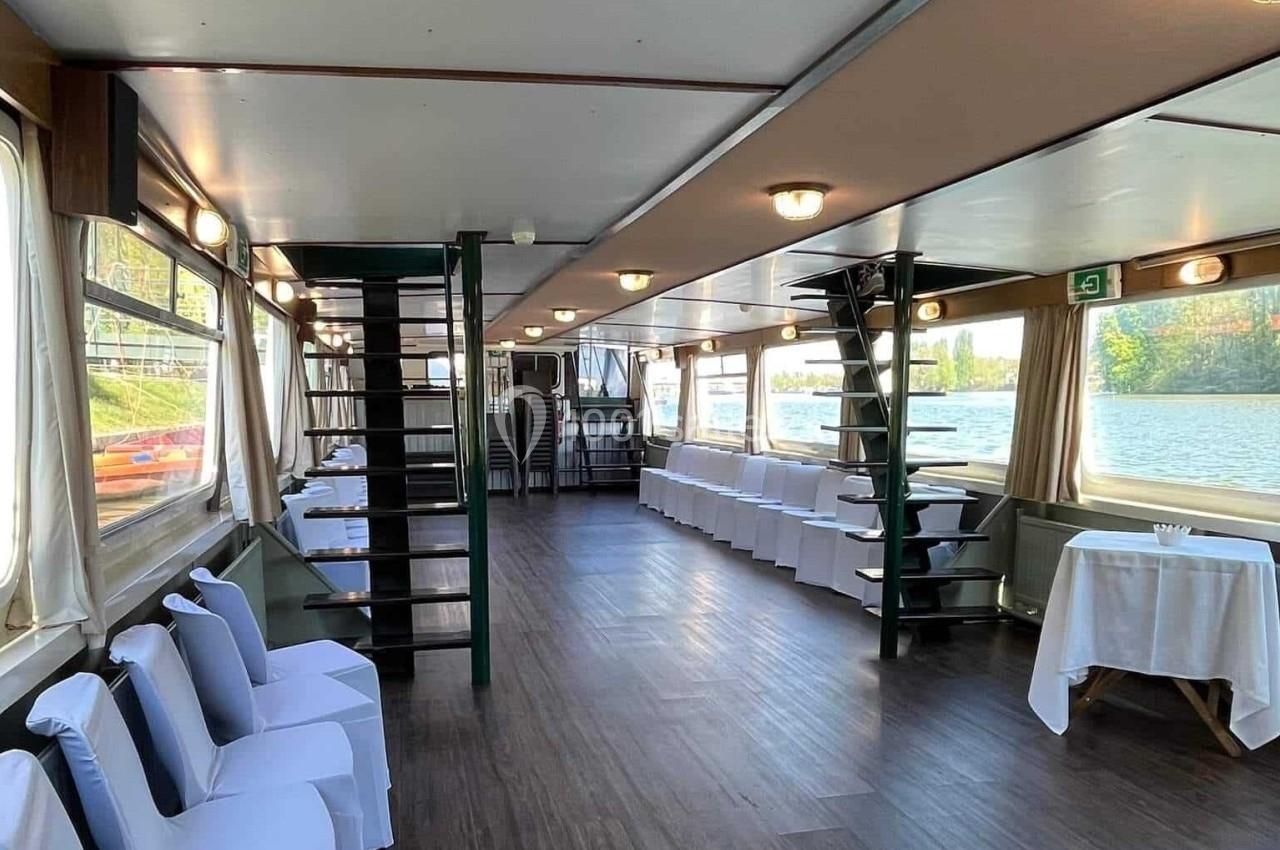 Salle de réception lumineuse sur un bateau, avec chaises blanches alignées et vue sur l'eau par de grandes fenêtres.