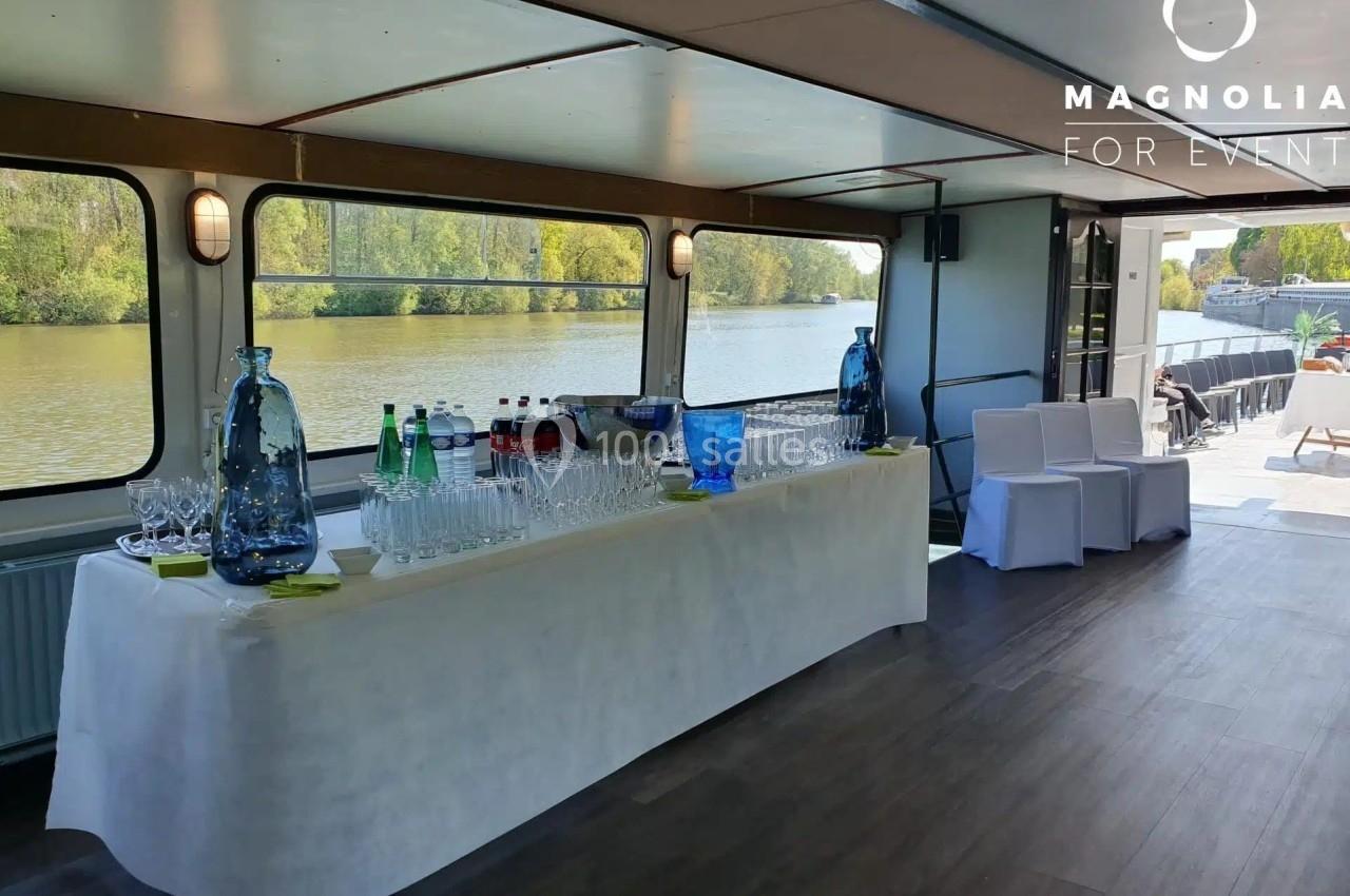 Table dressée avec des verres, bouteilles et carafes sur un bateau, vue sur un fleuve et des arbres à travers les fenêtres.