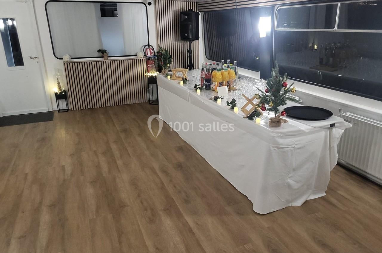 Salle avec parquet, table dressée avec boissons et décorations festives, éclairage tamisé et grandes fenêtres.