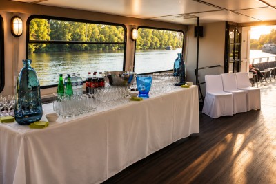 Intérieur d'une péniche avec un bar en bois, des chaises noires, des plantes et une vue sur l'eau par les fenêtres.