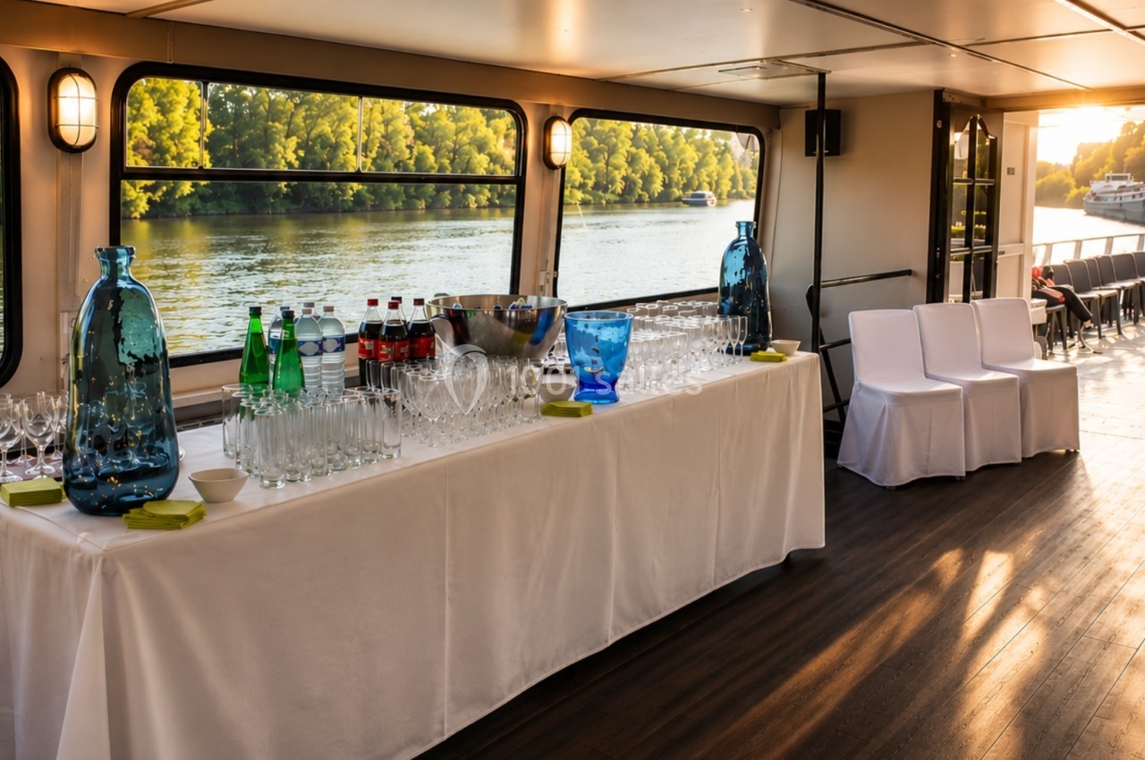 Table dressée avec verres, bouteilles et carafes sur un bateau, vue sur une rivière et arbres au coucher du soleil.