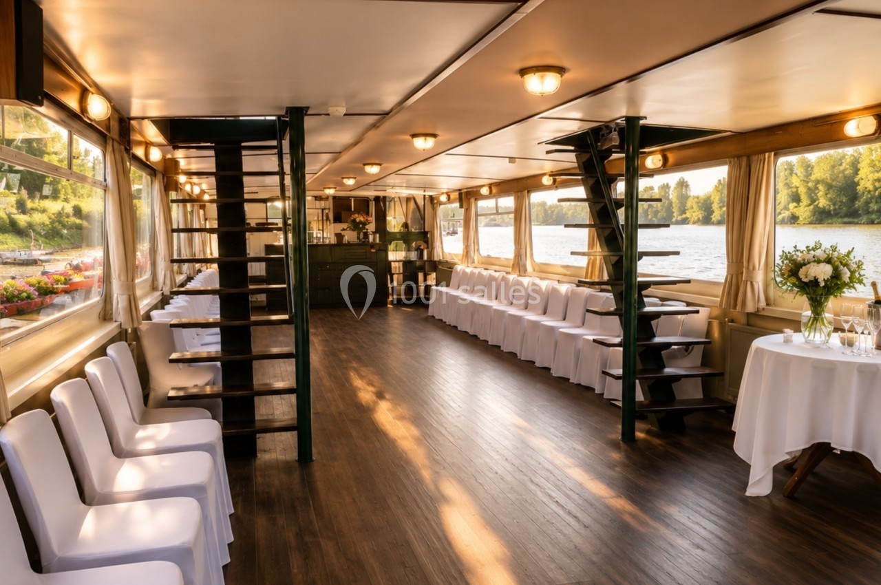 Salle intérieure d'un bateau avec des chaises blanches alignées, tables dressées et vue sur un paysage fluvial verdoyant.