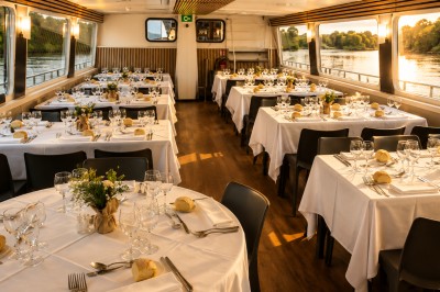 Intérieur d'une péniche avec un bar en bois, des chaises noires, des plantes et une vue sur l'eau par les fenêtres.