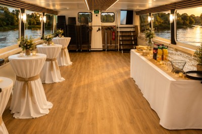 Intérieur d'une péniche avec un bar en bois, des chaises noires, des plantes et une vue sur l'eau par les fenêtres.