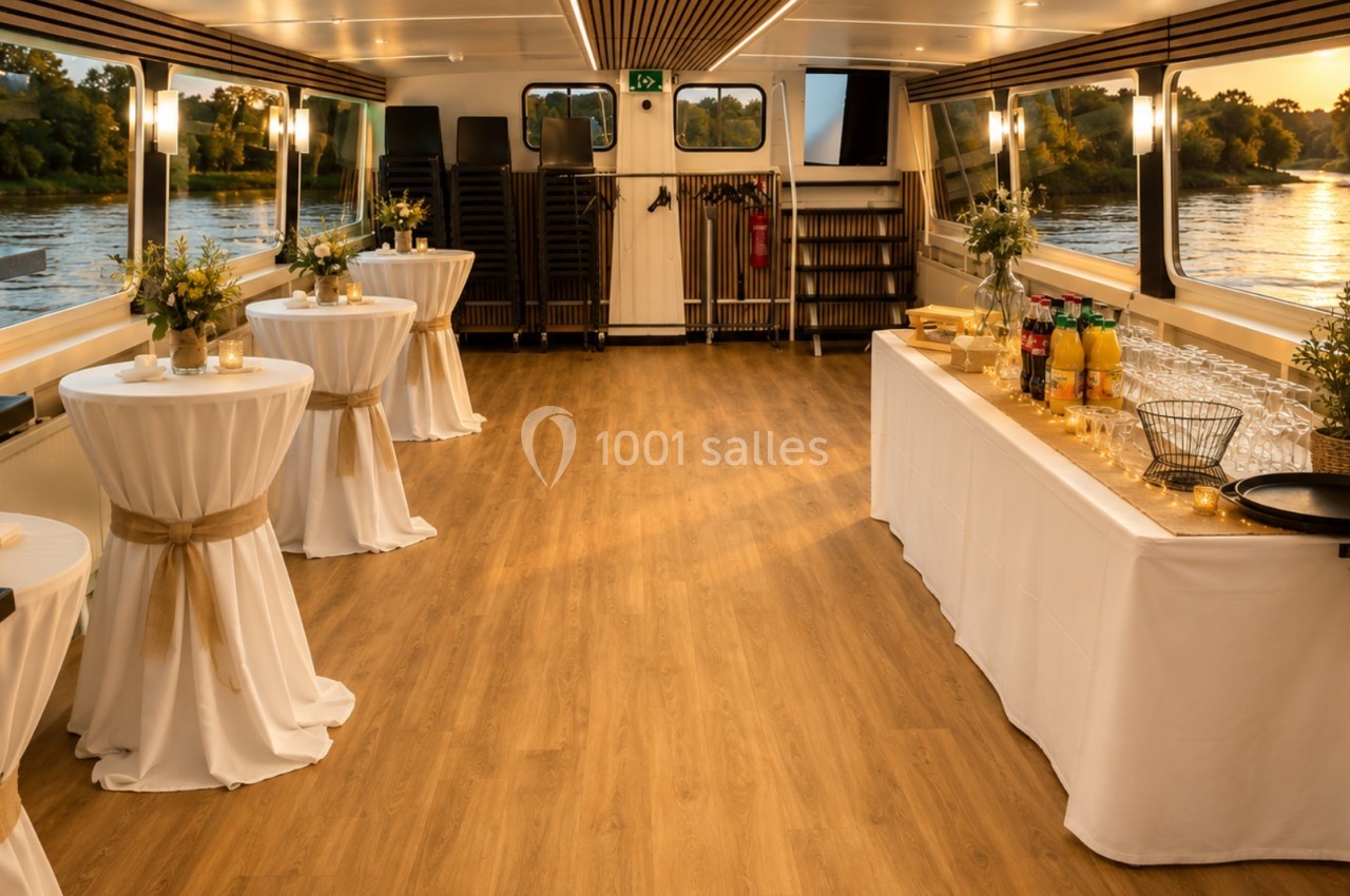 Salle de réception sur un bateau, avec tables hautes décorées et buffet installé, vue sur un fleuve à travers les fenêtres.