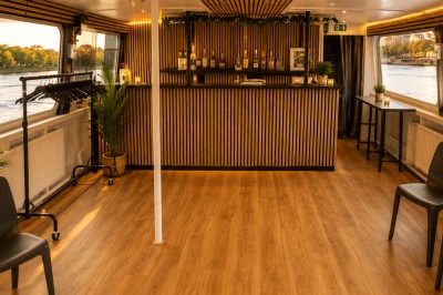 Intérieur d'une péniche avec un bar en bois, des chaises noires, des plantes et une vue sur l'eau par les fenêtres.