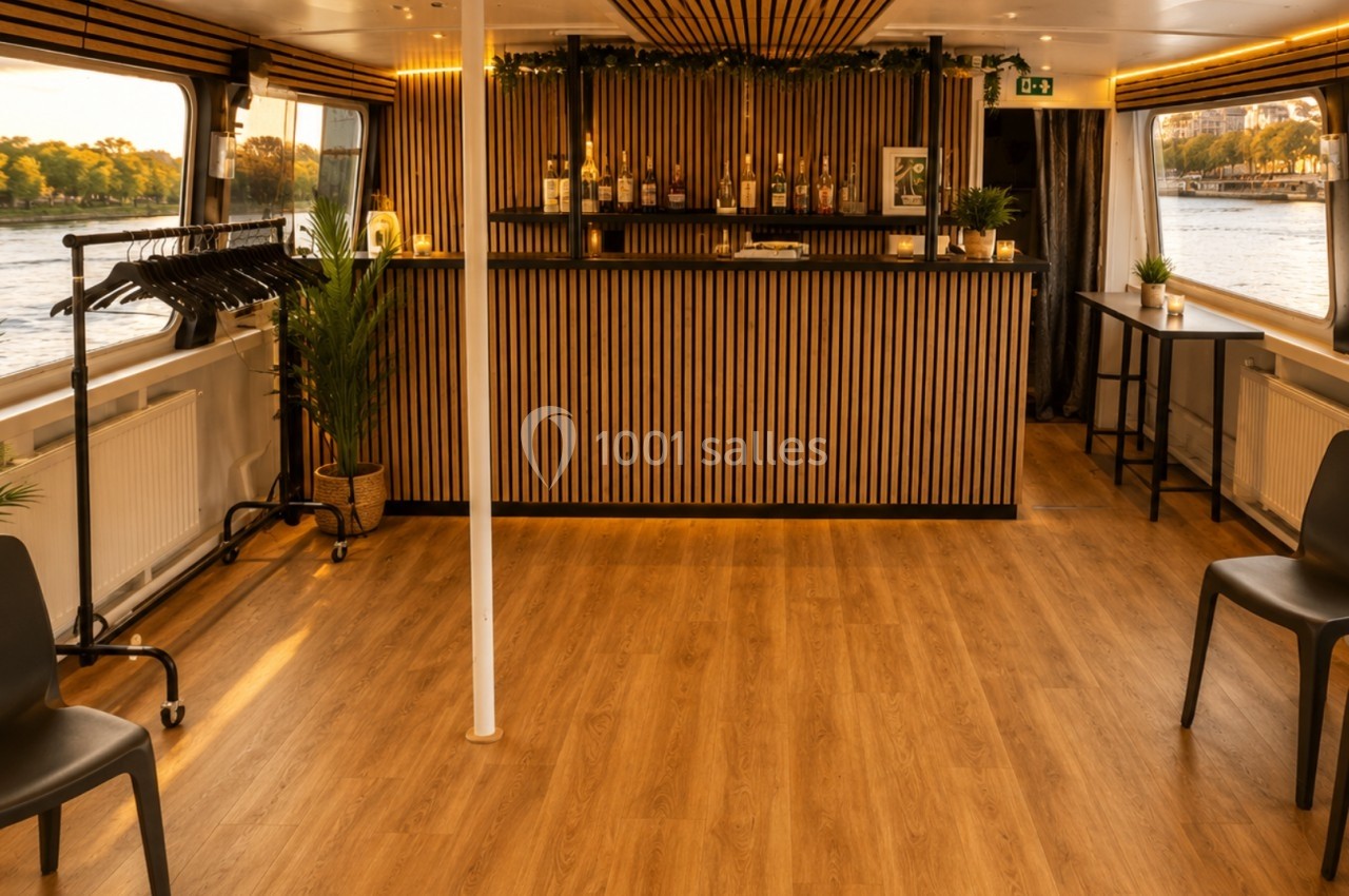 Intérieur d'une péniche avec un bar en bois, des chaises noires, des plantes et une vue sur l'eau par les fenêtres.