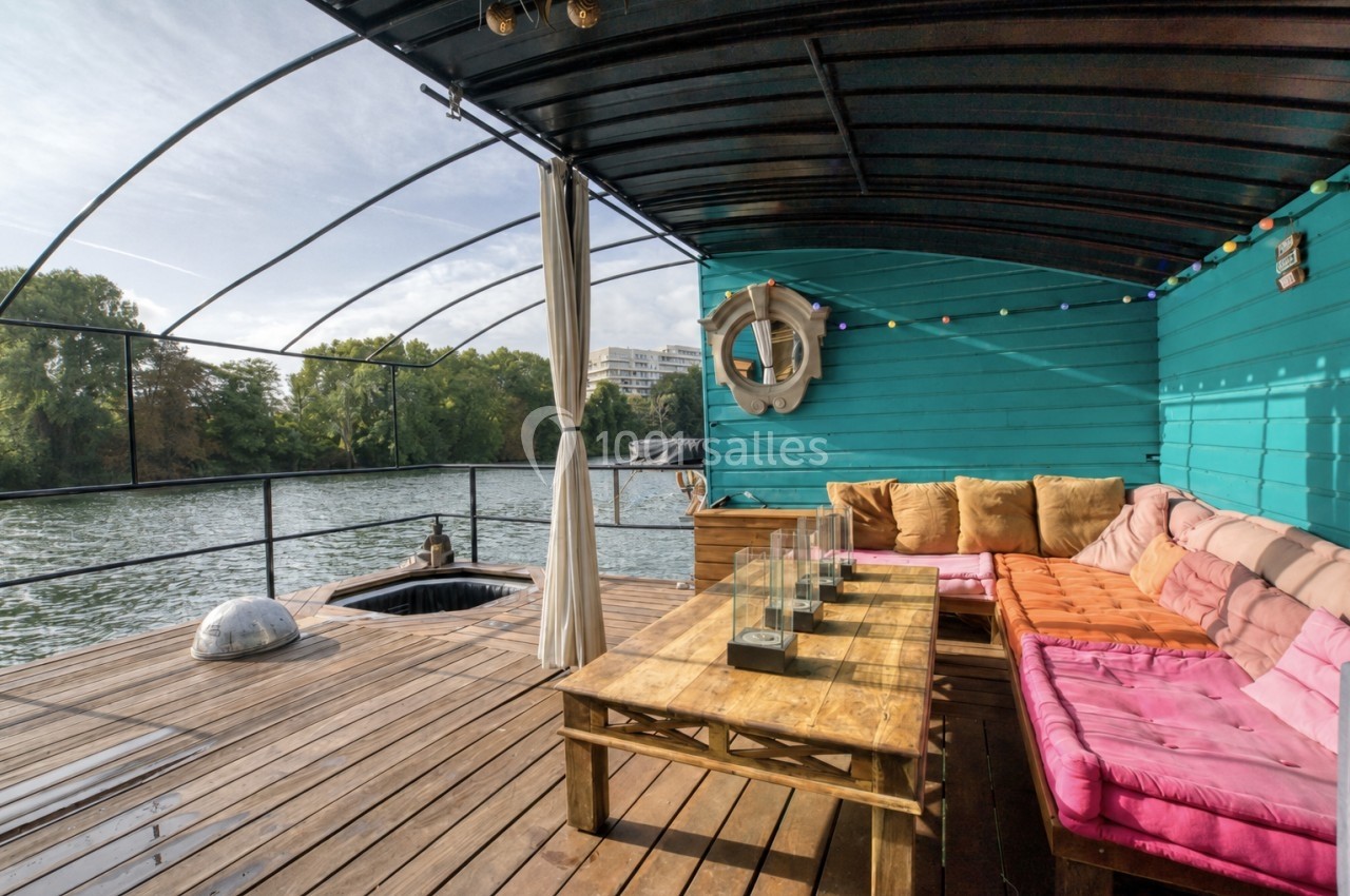 Terrasse en bois sur une péniche avec canapé coloré, table basse et vue sur une rivière bordée de végétation.