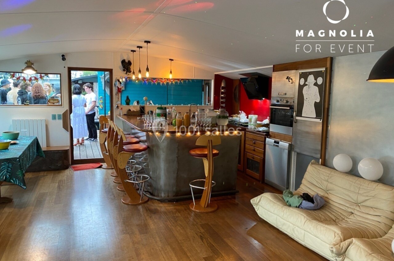 Intérieur d'une salle avec bar, tabourets, canapé, cuisine équipée et vue sur une terrasse avec des invités.