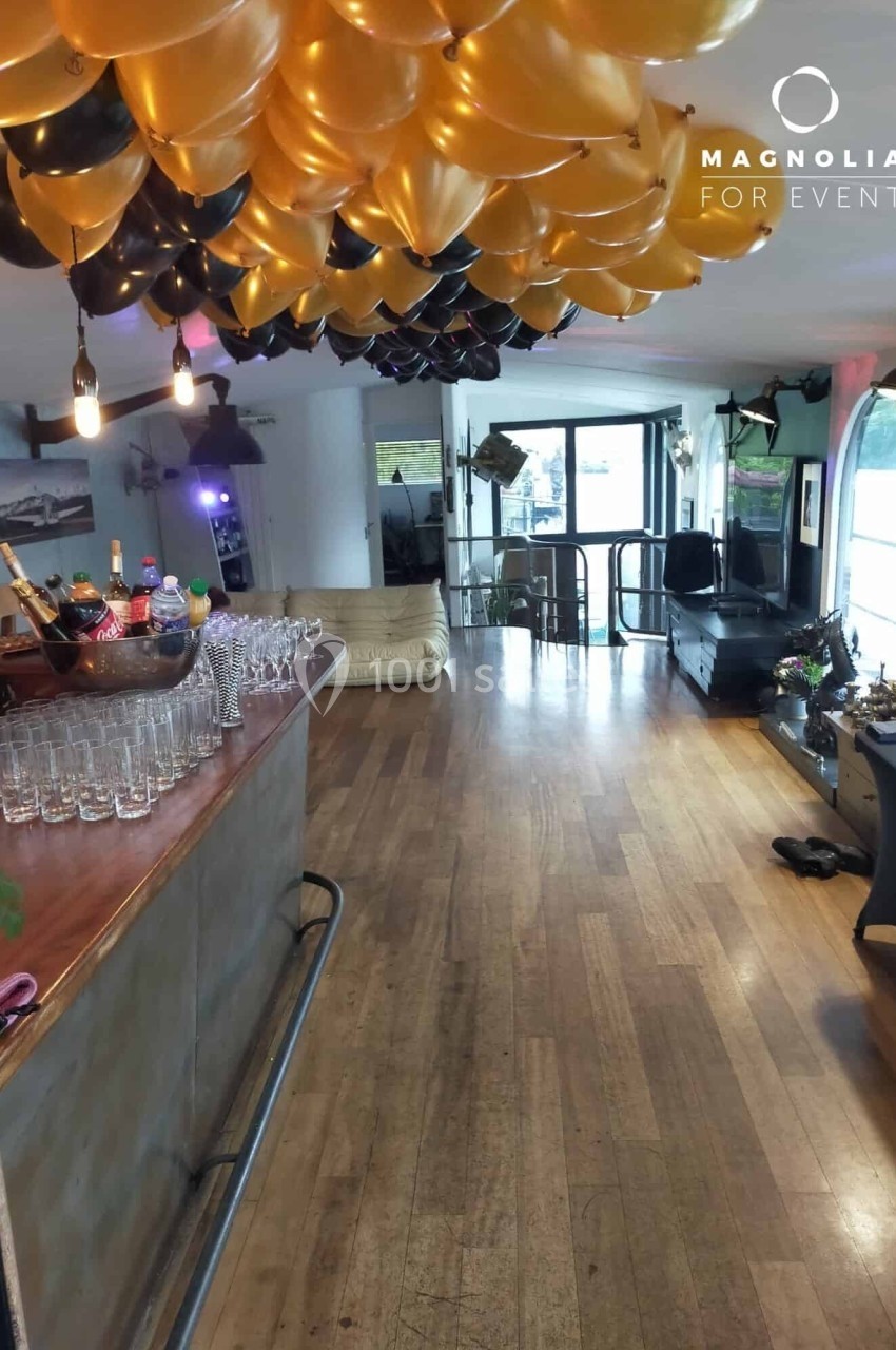 Salle décorée pour un événement avec des ballons dorés et noirs au plafond, bar équipé de verres et bouteilles.