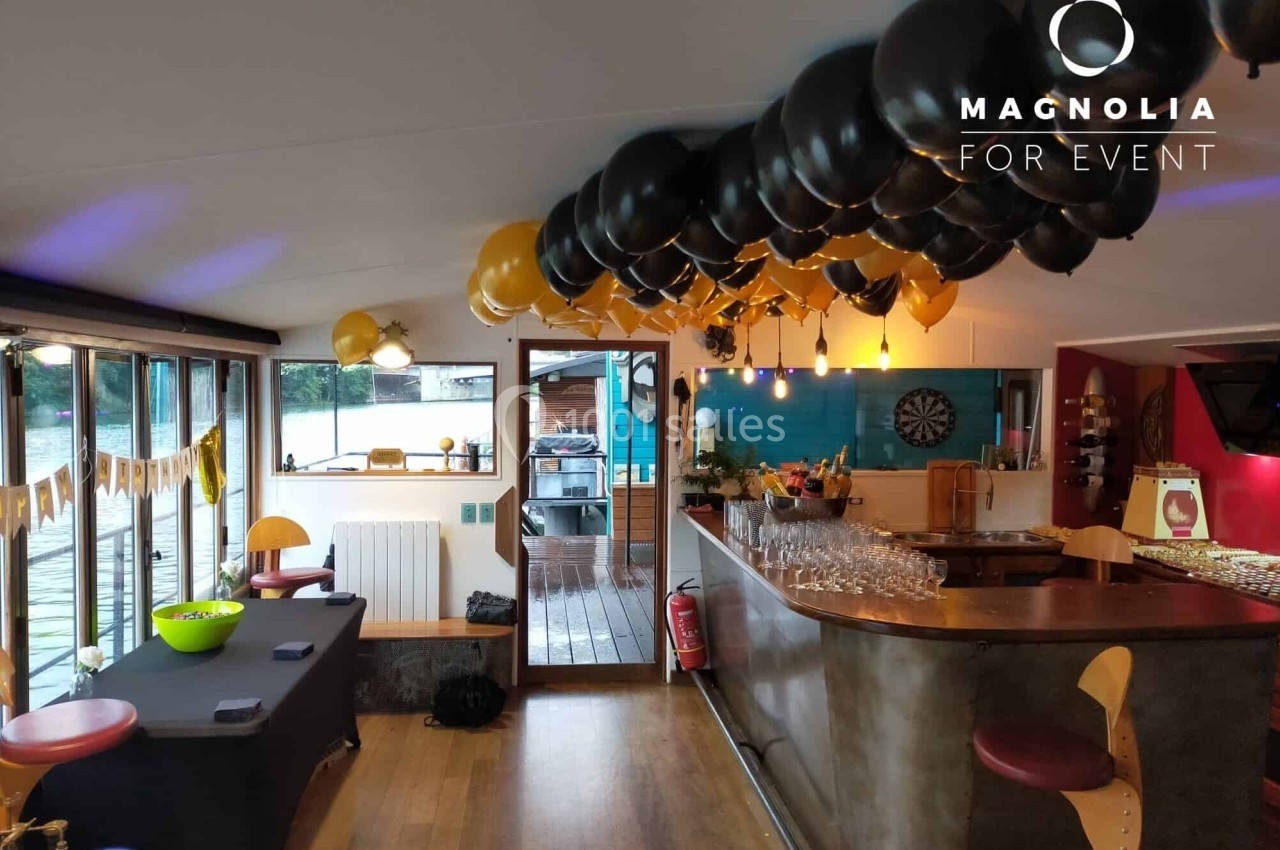 Salle décorée pour un événement avec ballons noirs et dorés, bar équipé et vue sur une terrasse extérieure.