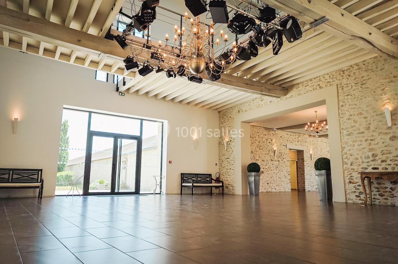 Salle spacieuse avec murs en pierre, sol carrelé, plafond en poutres apparentes et lustres suspendus.